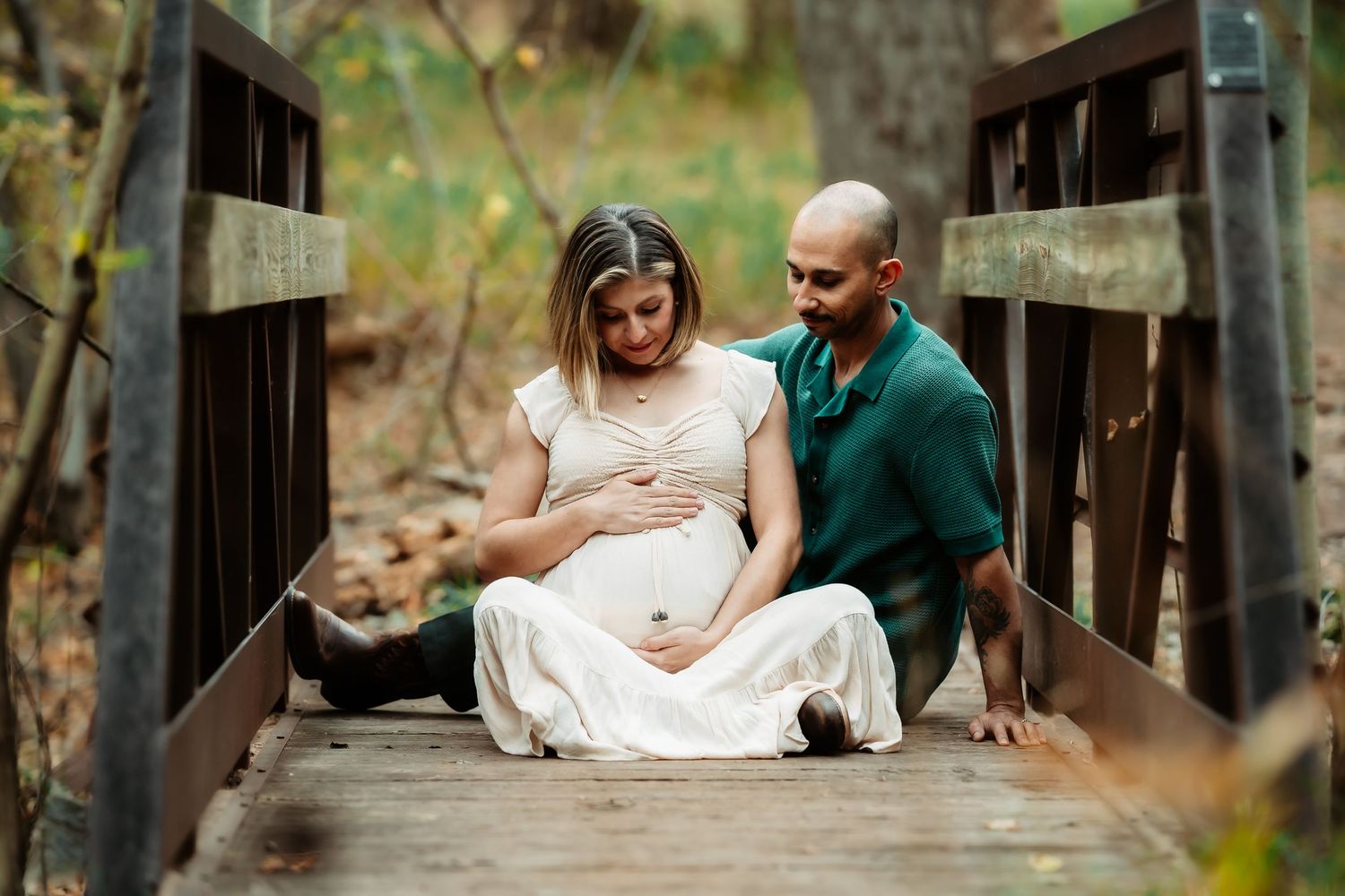 A couple shares an intimate moment while sitting together on a wooden footbridge in a park.