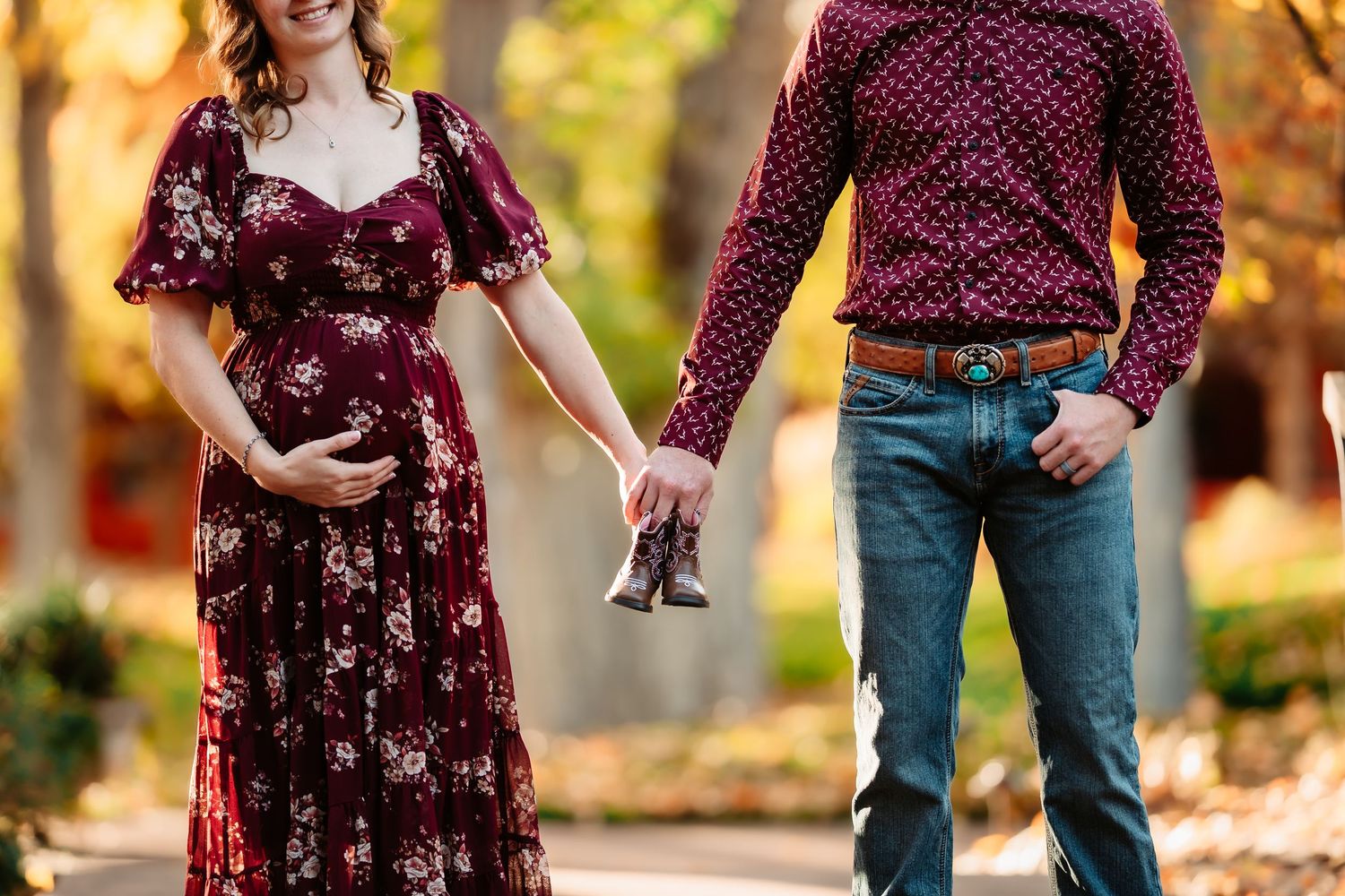 A couple in burgundy floral dress and denim poses outdoors during autumn with golden sunlight filtering through trees.