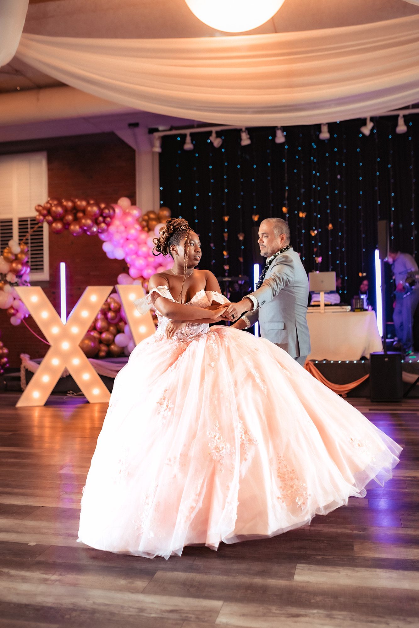 Elegant couple dances at a wedding reception with twinkle lights and pink balloons decorating the venue.