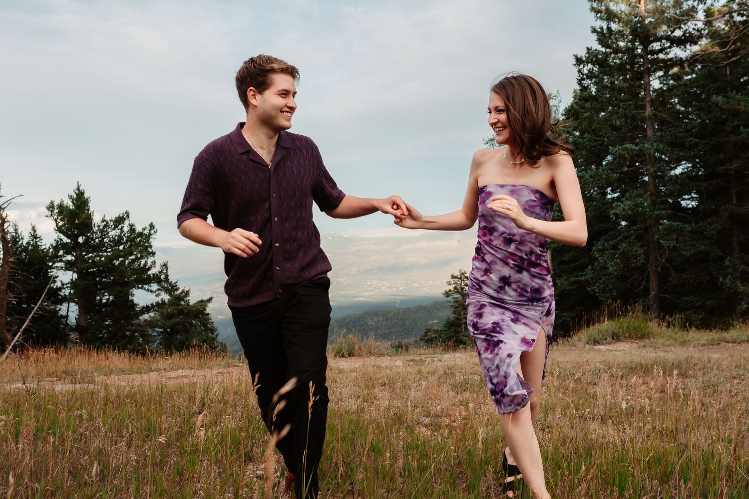Two people run playfully through a mountain meadow wearing purple and black outfits.
