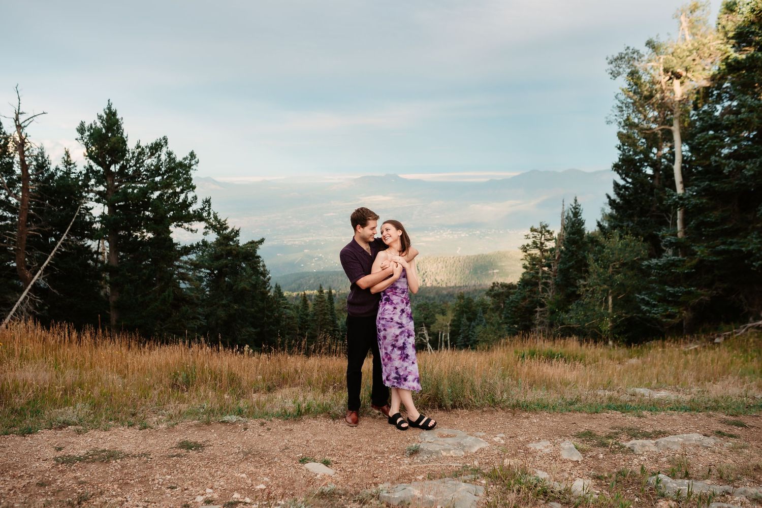 Wide landscape view of couple embracing on mountaintop during sunset engagement session.