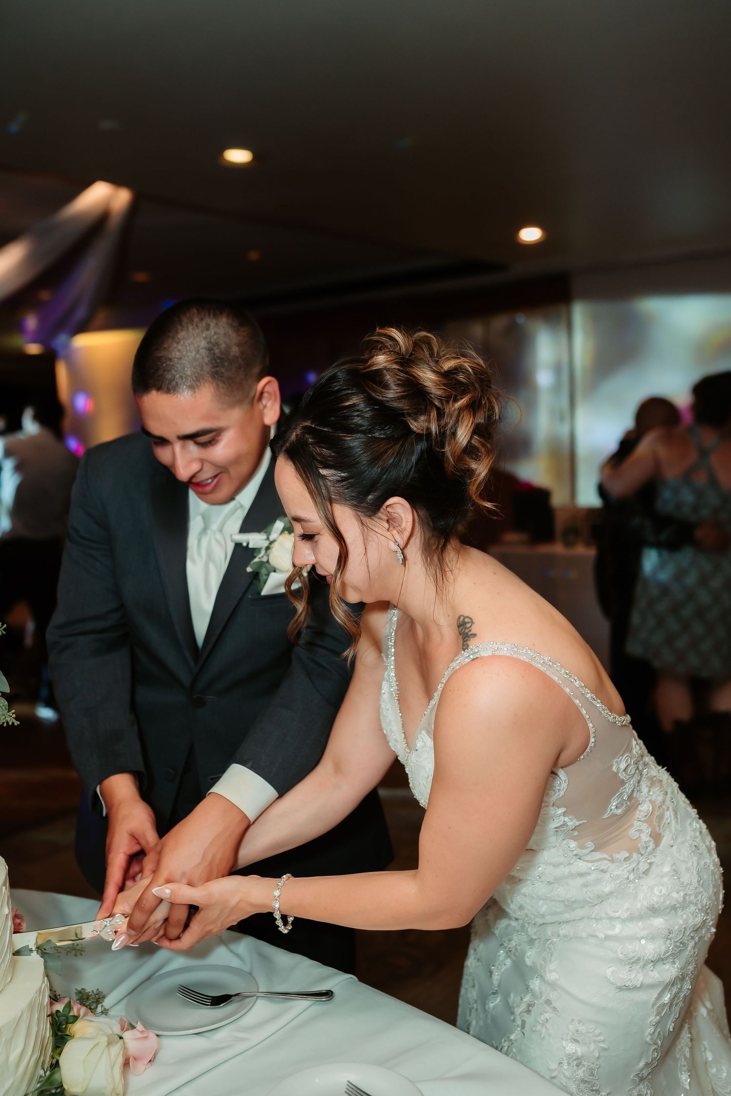 A couple in formal wedding attire smiling while cutting their wedding cake together at their reception.