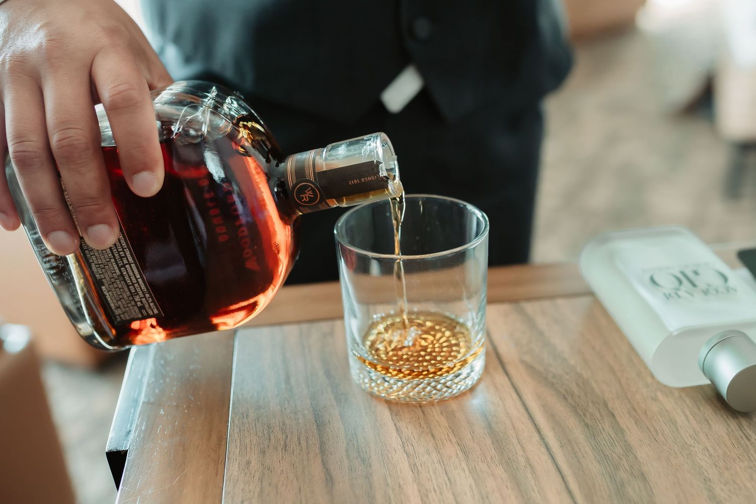 A person pours amber liquor into a glass at a bar with several bottles in the background.