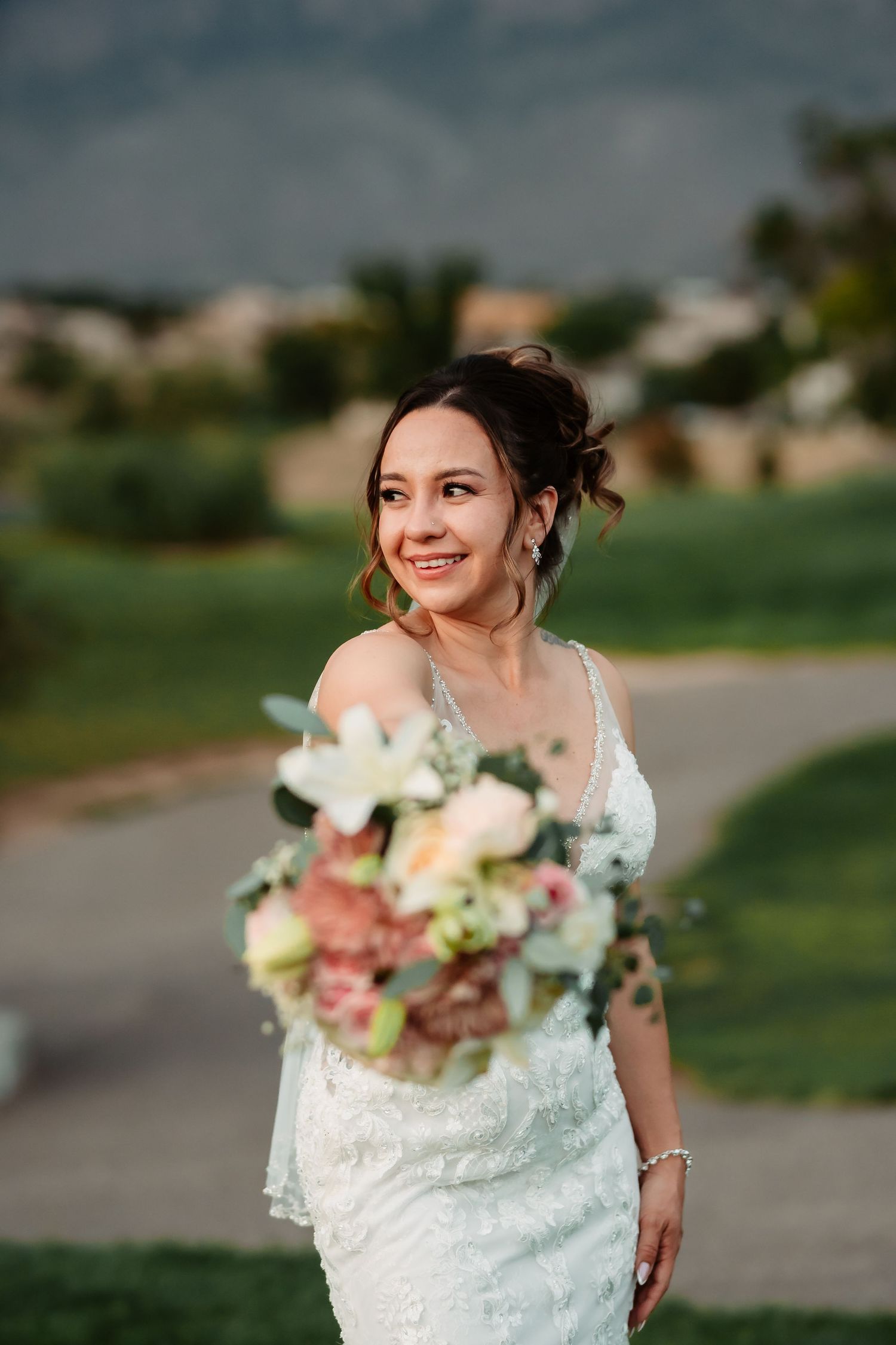 A bride in a white lace dress holds a pastel bouquet while posing outdoors on a grassy hill.