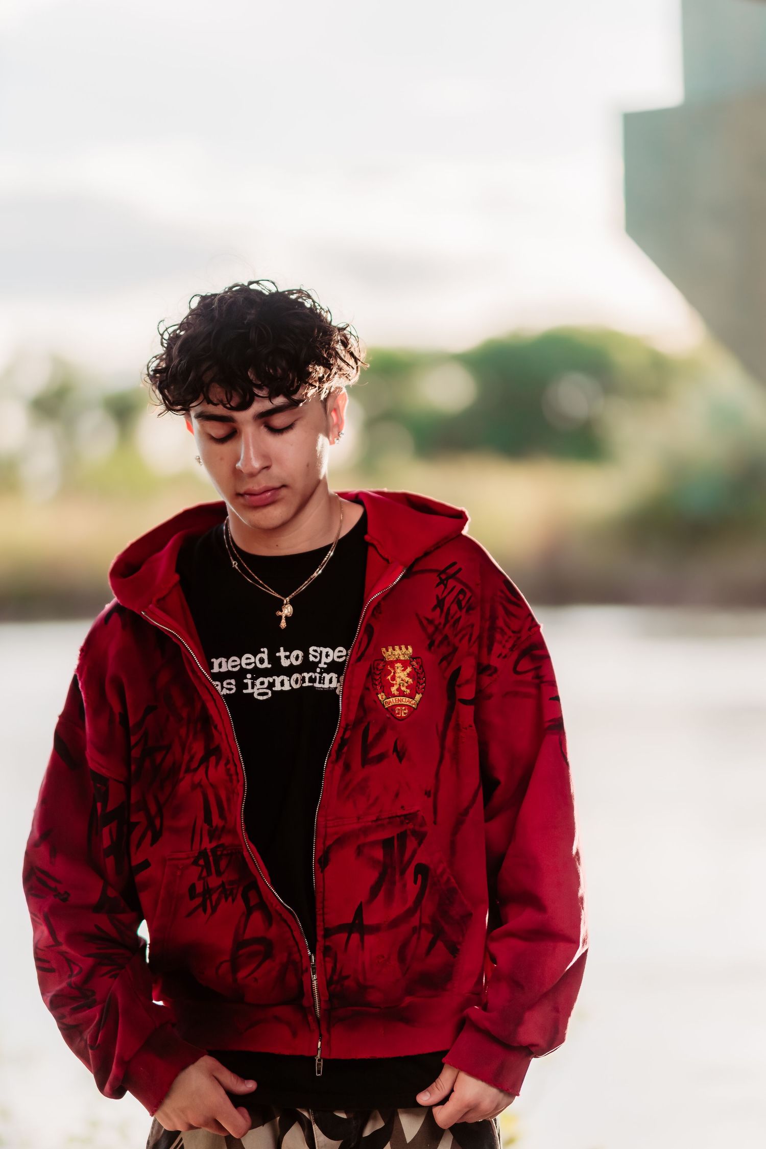 Young model poses outdoors wearing a red patterned hoodie and black t-shirt in natural lighting.