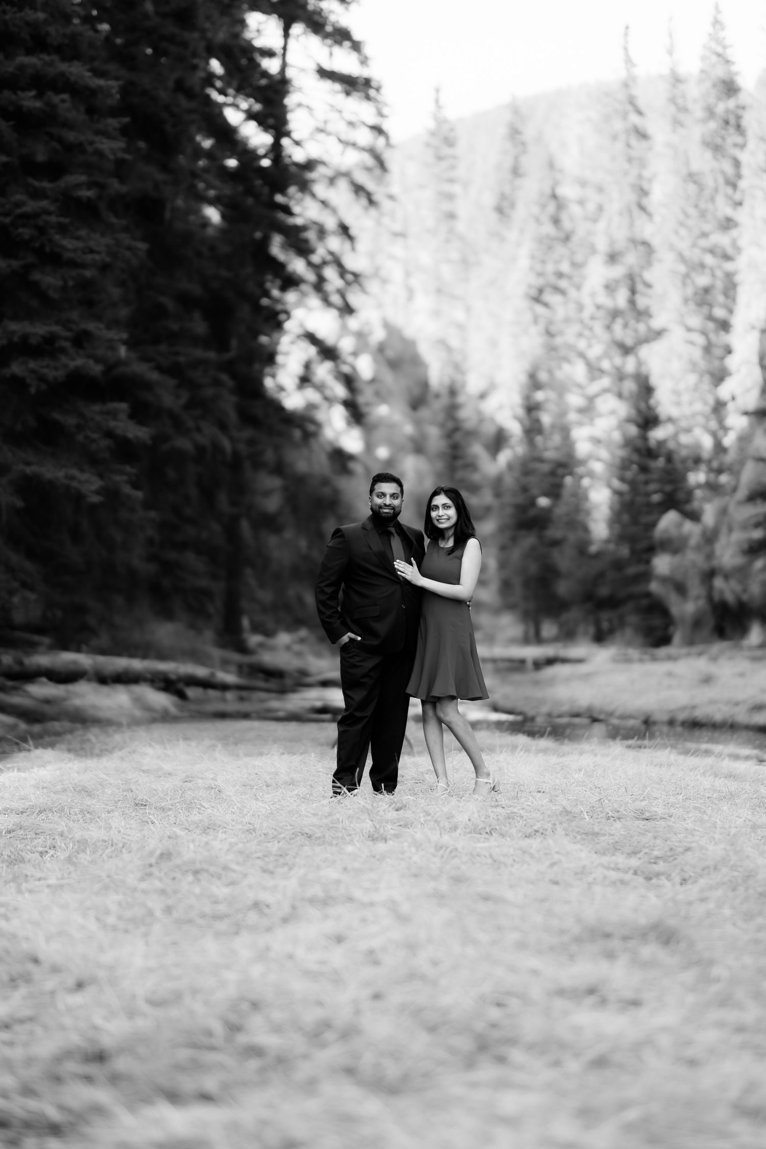 A black and white portrait of a couple standing in a mountain meadow surrounded by evergreen trees.