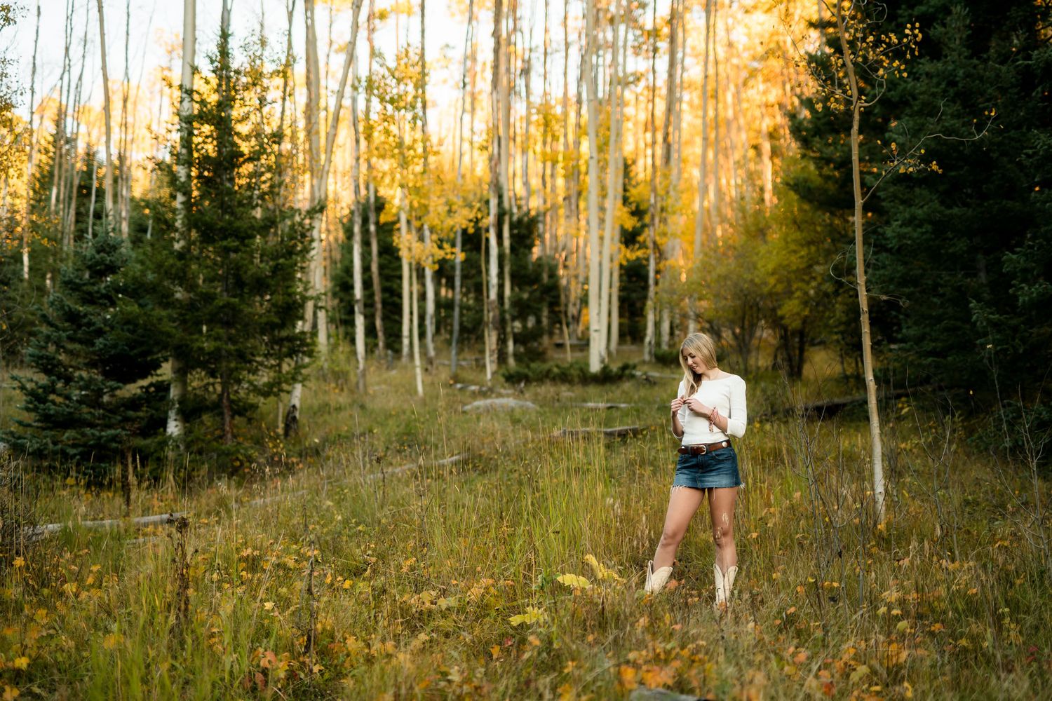 A young woman in a white top and denim shorts stands in a golden aspen forest during autumn.