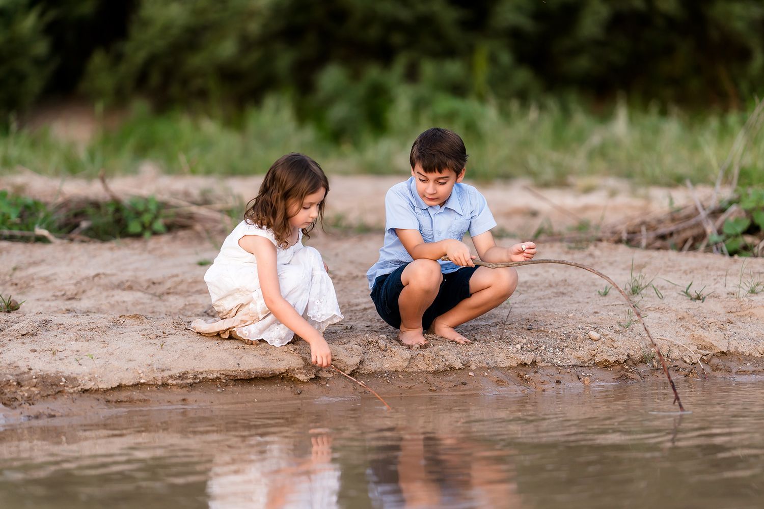 Two young children play together in the sand near a calm river on a sunny day.