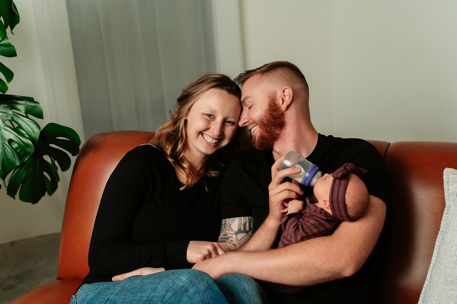 Happy couple sitting on a brown leather sofa, smiling together while holding their newborn baby wrapped in purple.