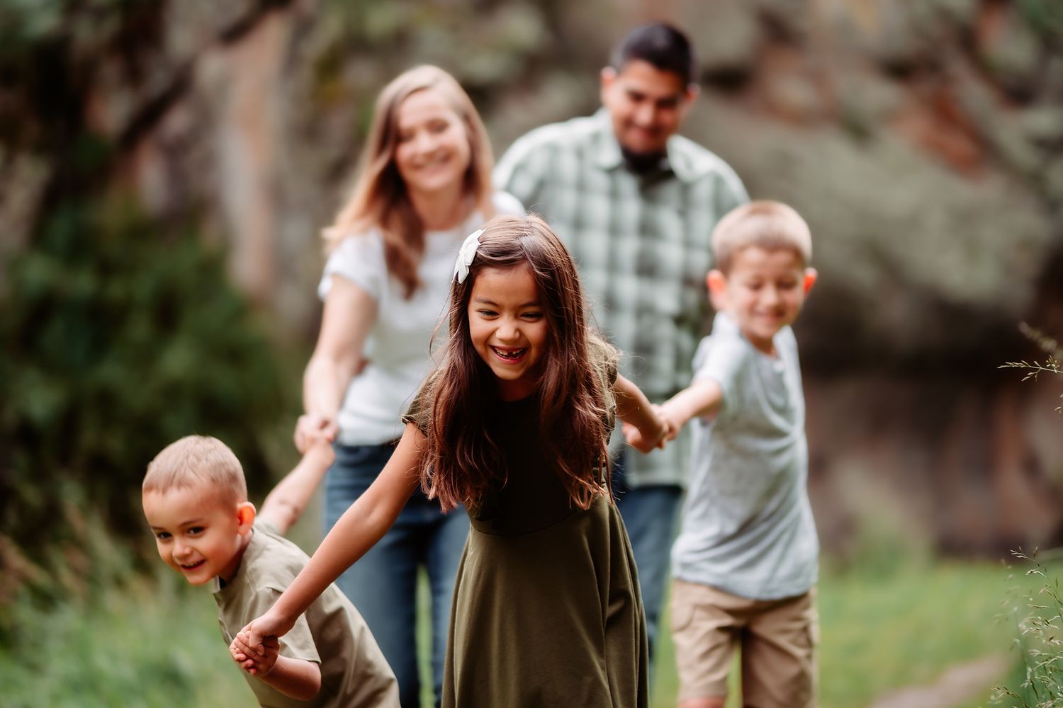 Happy family of five walking outdoors, parents smiling as three young children lead the way through a lush green park.