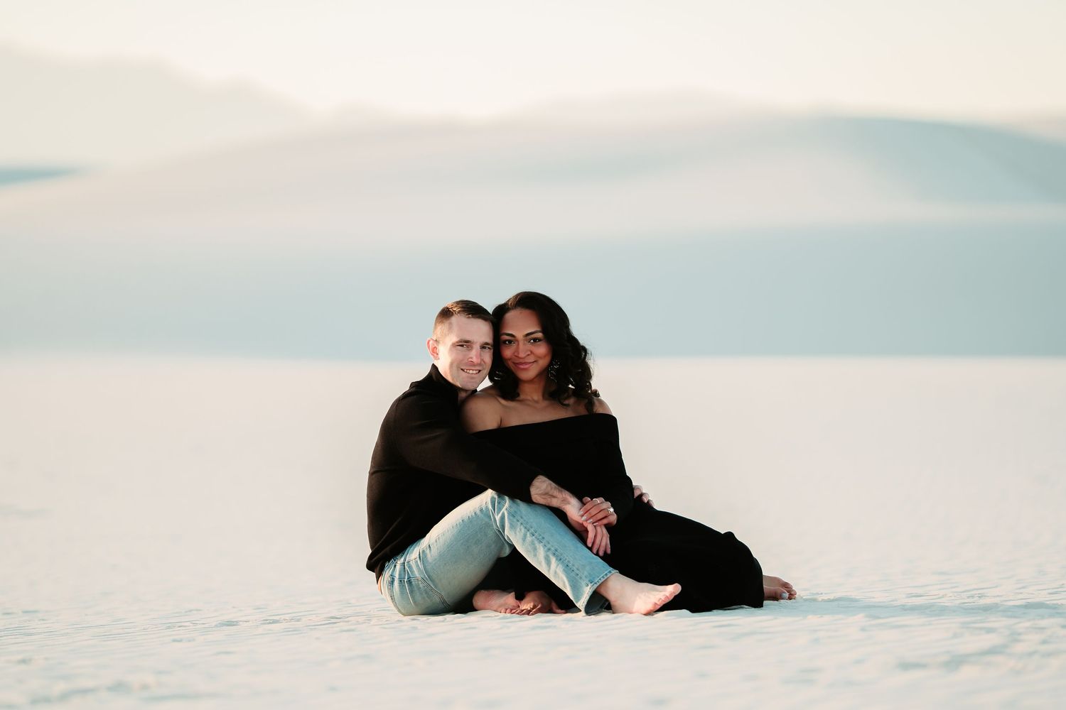 A couple sits close together on white sand during sunset with a pastel sky in the background.