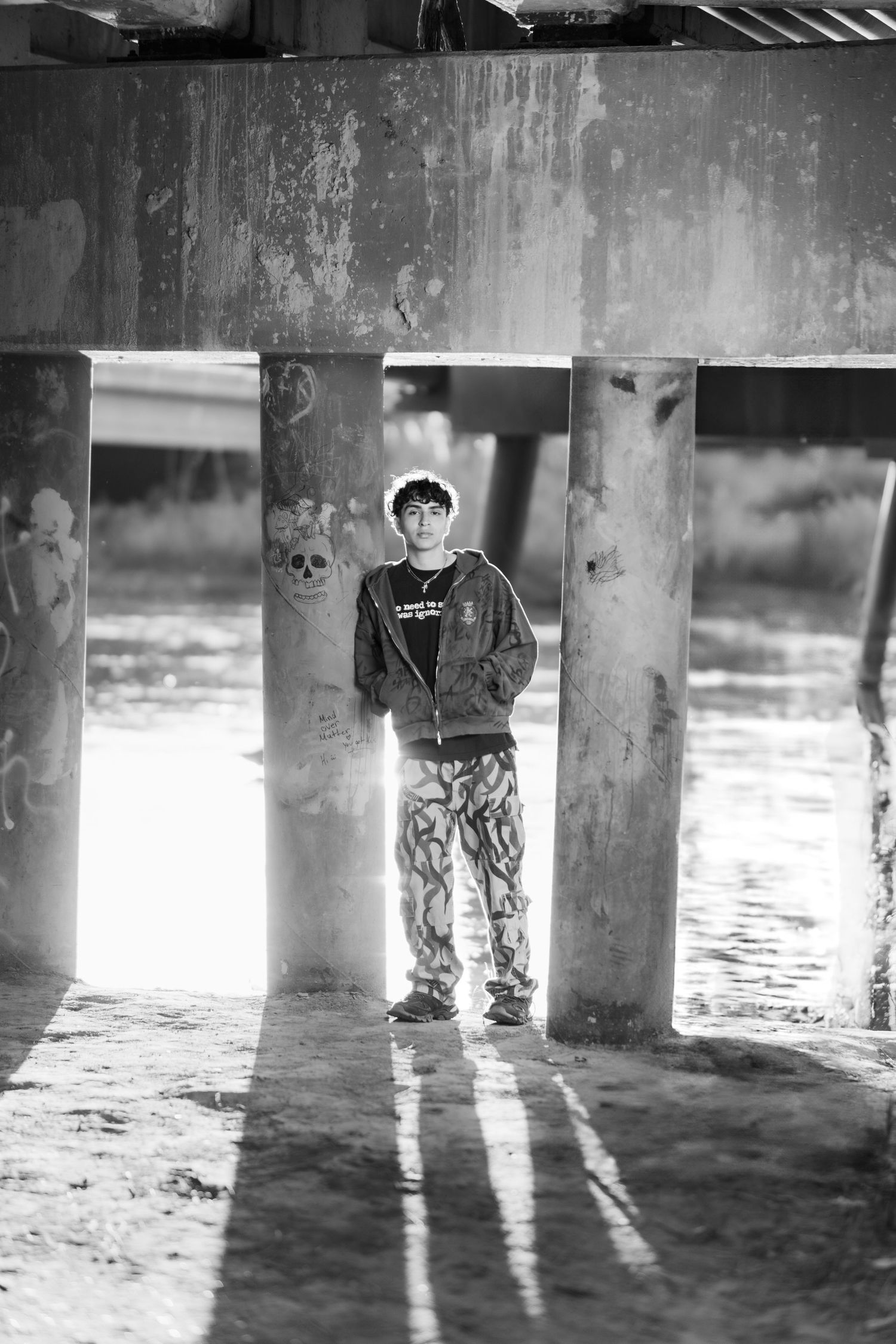 A black and white photo of someone standing between concrete pillars under a bridge with water visible in the background.
