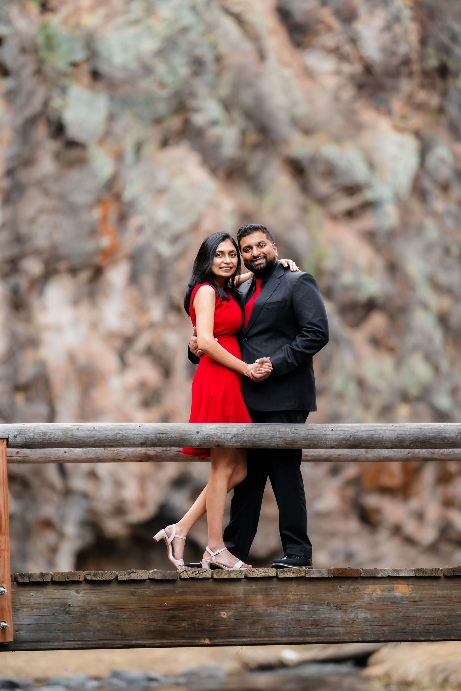 A couple in formal attire poses romantically on a wooden bridge against a rocky cliff backdrop.