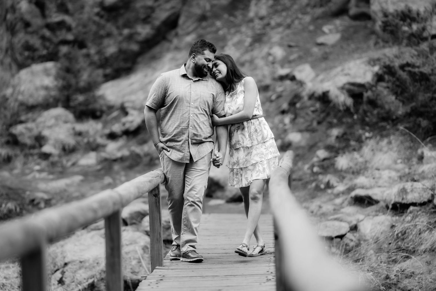 A couple shares an intimate moment while standing together on a wooden footbridge in a natural setting.