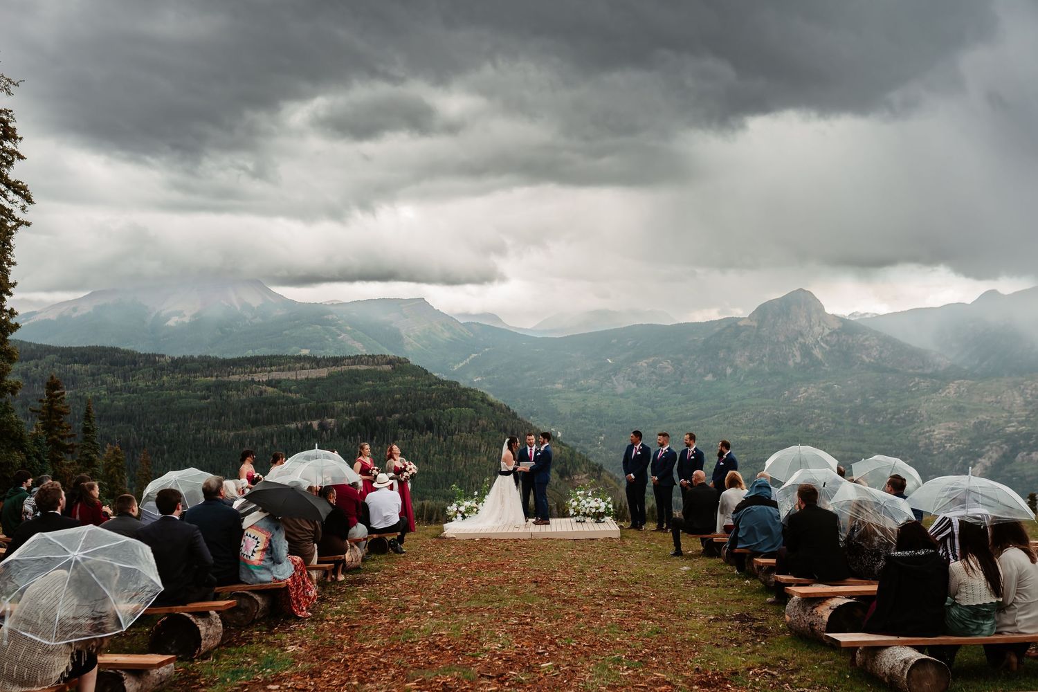 Scenic mountain ceremony setup with wooden benches and white umbrellas against dramatic cloudy skies and mountain peaks.