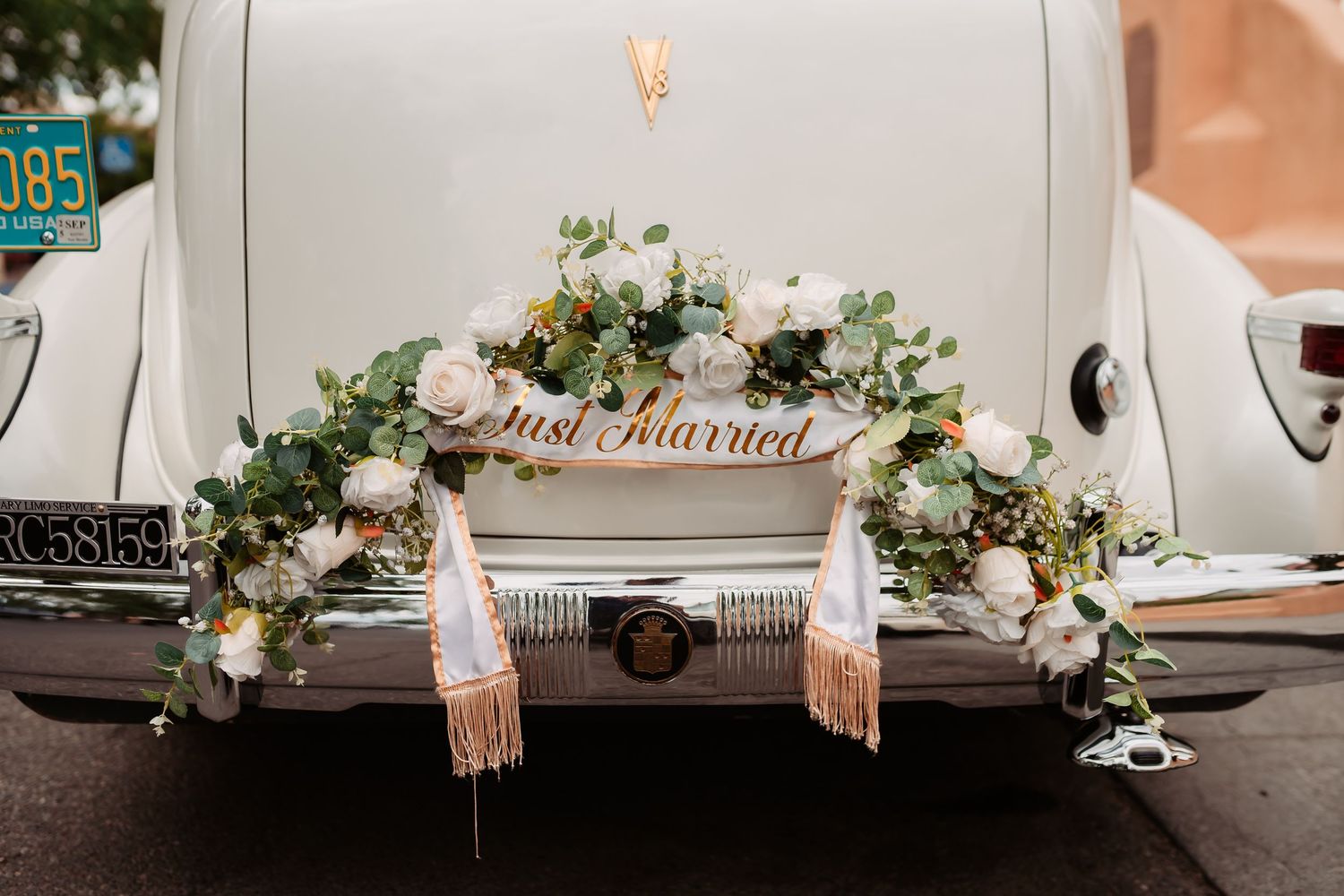 White vintage wedding car with delicate floral decorations and peachy ribbons parked outside a brick building.