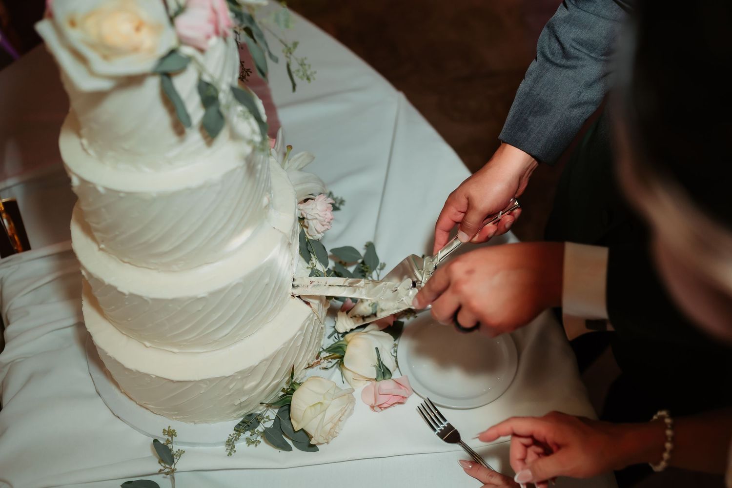 A tiered white wedding cake with floral decorations is being cut at a wedding reception.