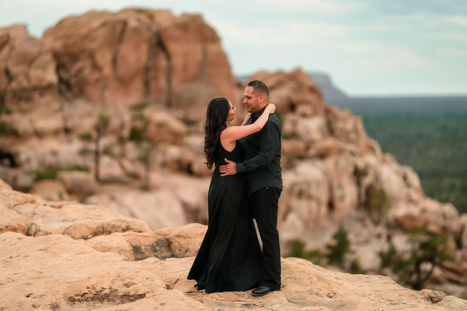 Couple embracing on desert rock landscape in matching black outfits with red rock formations behind.