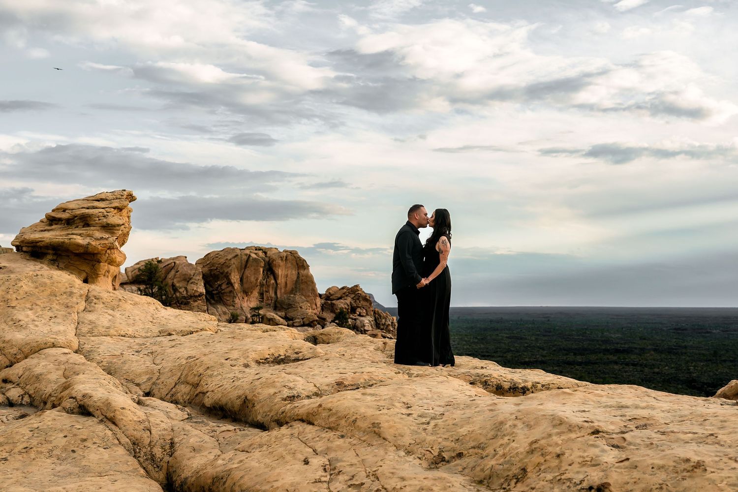 Couple kissing in black outfits on sandstone rock formation with expansive horizon and dramatic clouds.