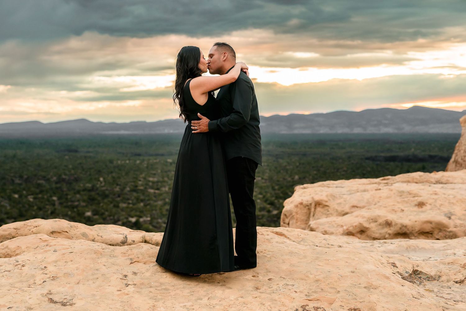 Couple in black formalwear kissing on rocky overlook with mountain valley sunset view.
