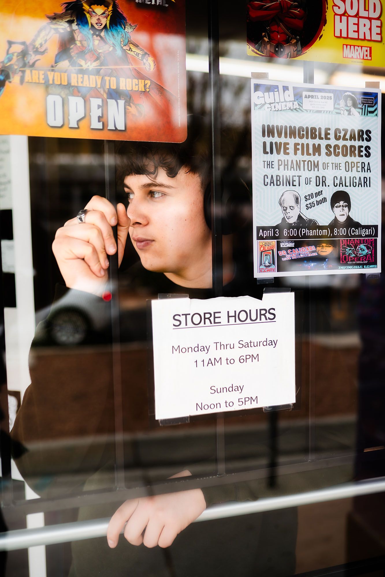 Young man peering through glass store door with open sign and posted store hours notice.