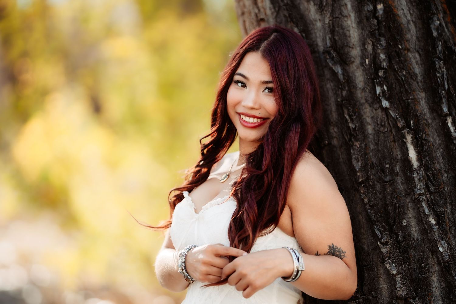 A young woman with long dark hair poses outdoors in front of autumn trees wearing a white sleeveless top.