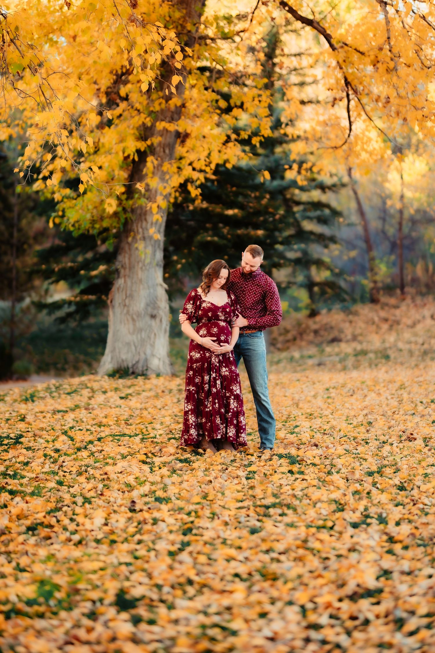 A couple stands together surrounded by golden autumn leaves and evergreen trees in a romantic outdoor setting.