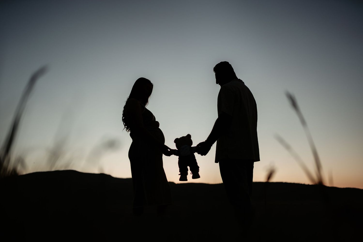 Silhouetted figures hold hands with small child against sunset sky in emotional family moment.