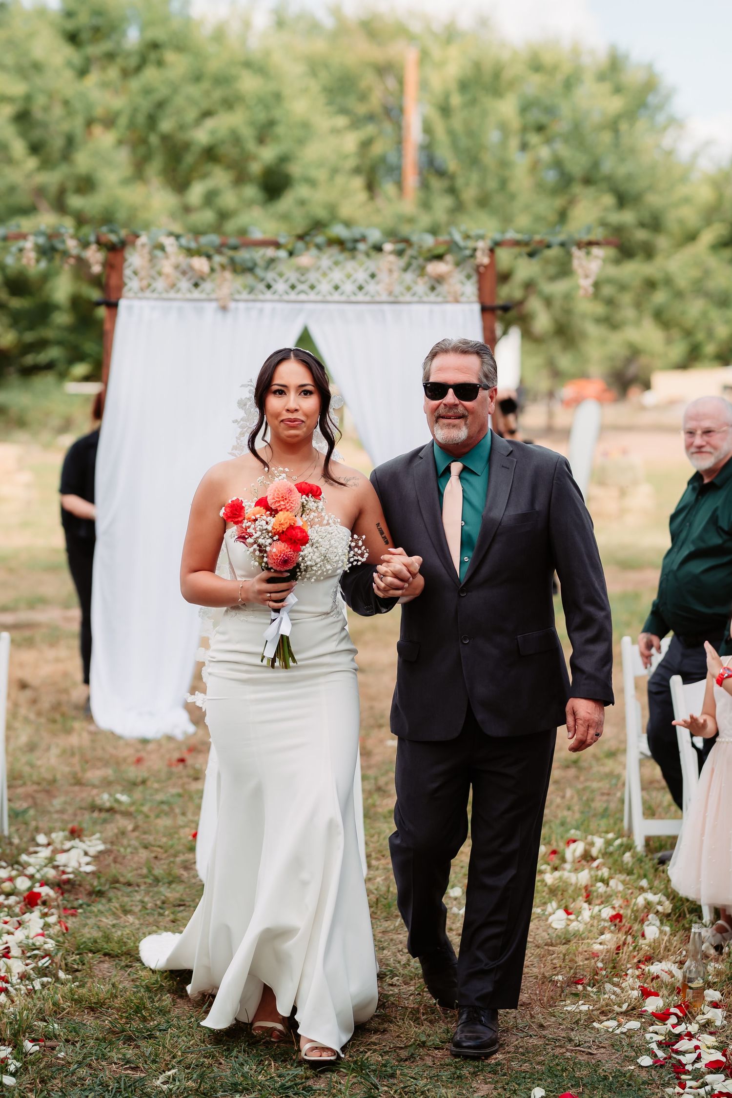 A wedding ceremony outdoors with a white backdrop, floral decorations, and rose petals scattered along the aisle.