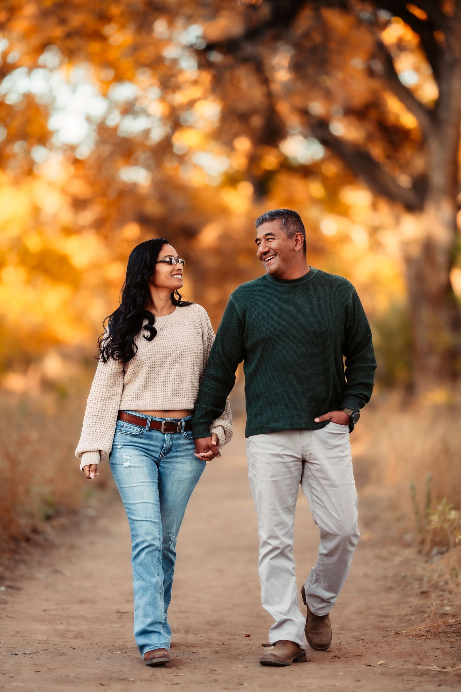 A couple walking hand in hand on a dirt path during autumn with golden sunlight filtering through trees in background.