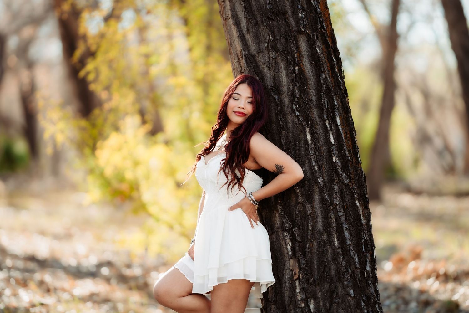 Woman in white dress poses outdoors in autumn forest setting during golden hour photography session.