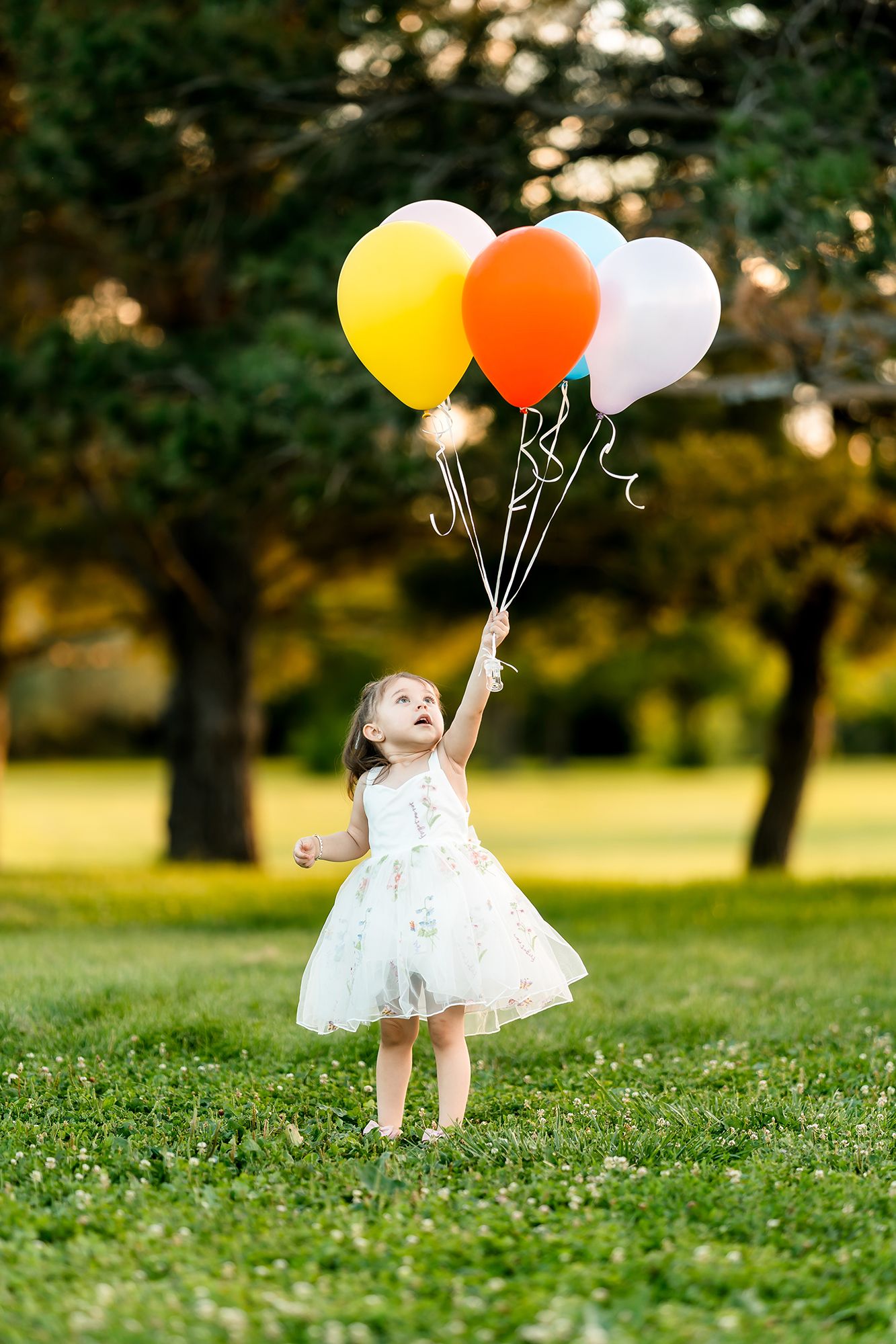 A joyful little girl in a white dress holds colorful balloons in a sunny green park.