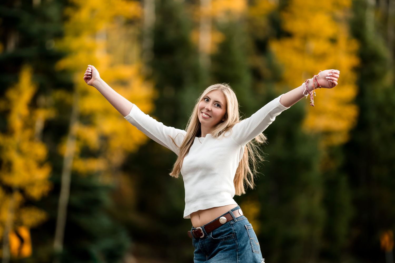 Joyful blonde woman in white crop top raising arms outdoors with colorful autumn trees in background.