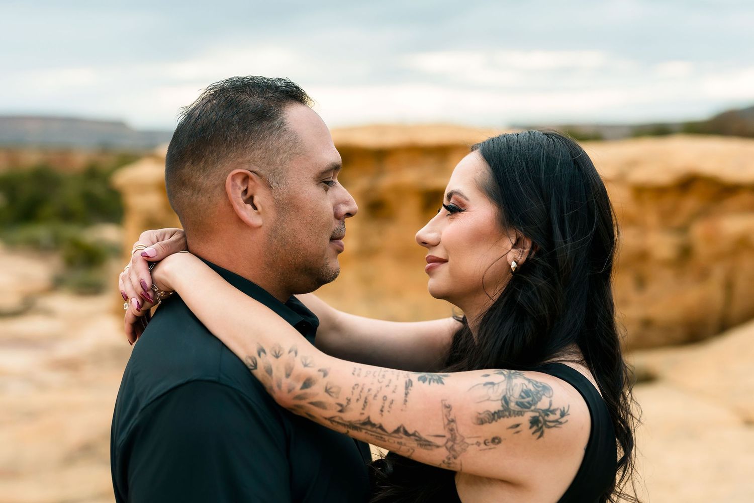 Tattooed woman embracing man face-to-face among warm sandstone rocks in desert setting.