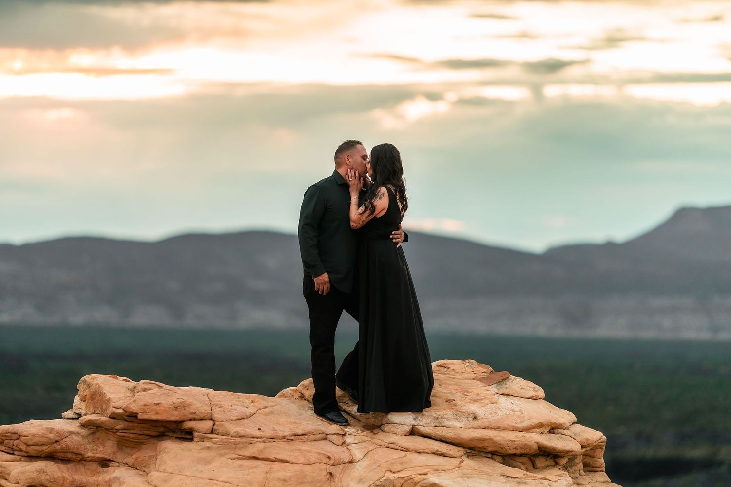 Couple in black attire sharing a kiss on a rocky cliff at sunset with mountain landscape backdrop.