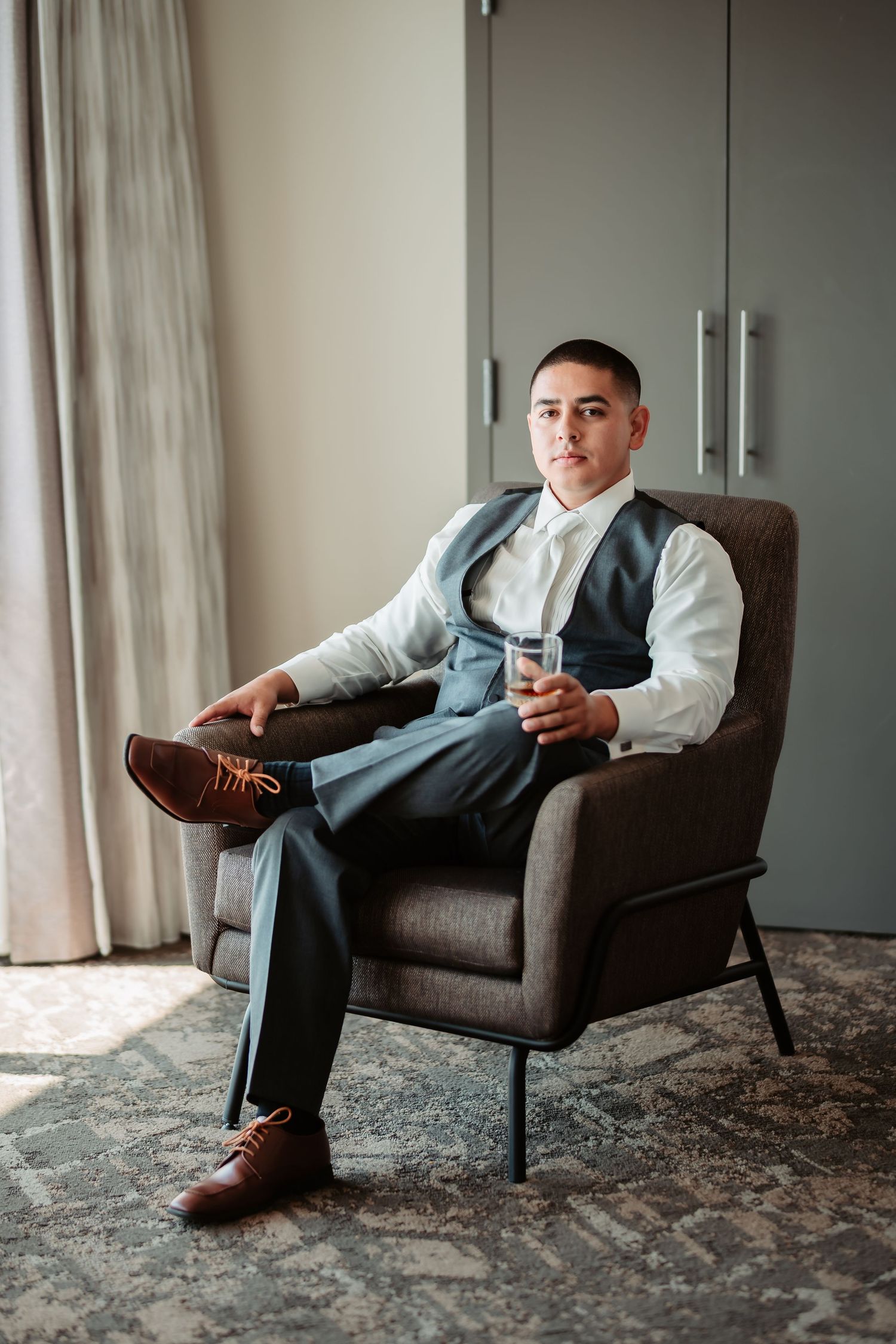 A person in formal attire sits in a gray armchair holding a glass, wearing a vest and brown dress shoes.