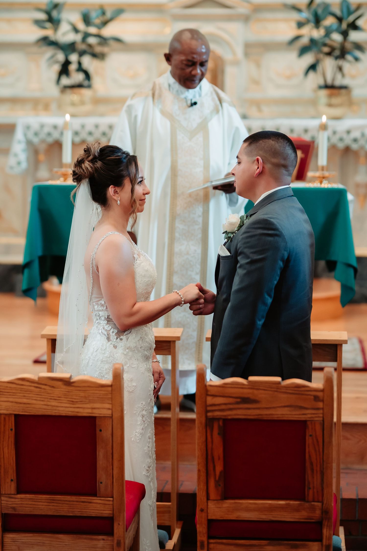 A couple shares a lighthearted moment during their wedding ceremony while seated in wooden chairs at a church.