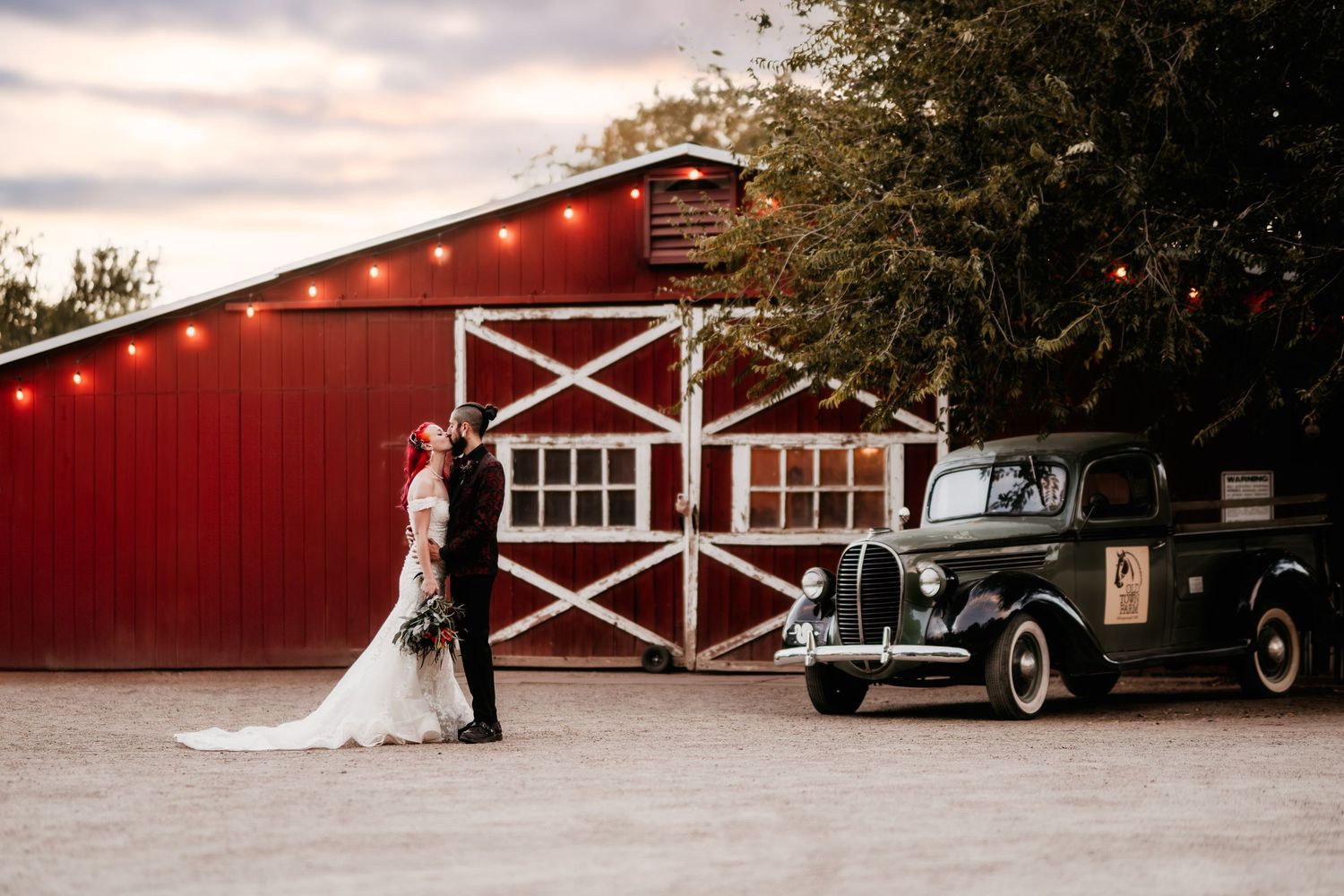 Vintage black car parked in front of a red rustic barn while a bride and groom embrace during a country wedding.