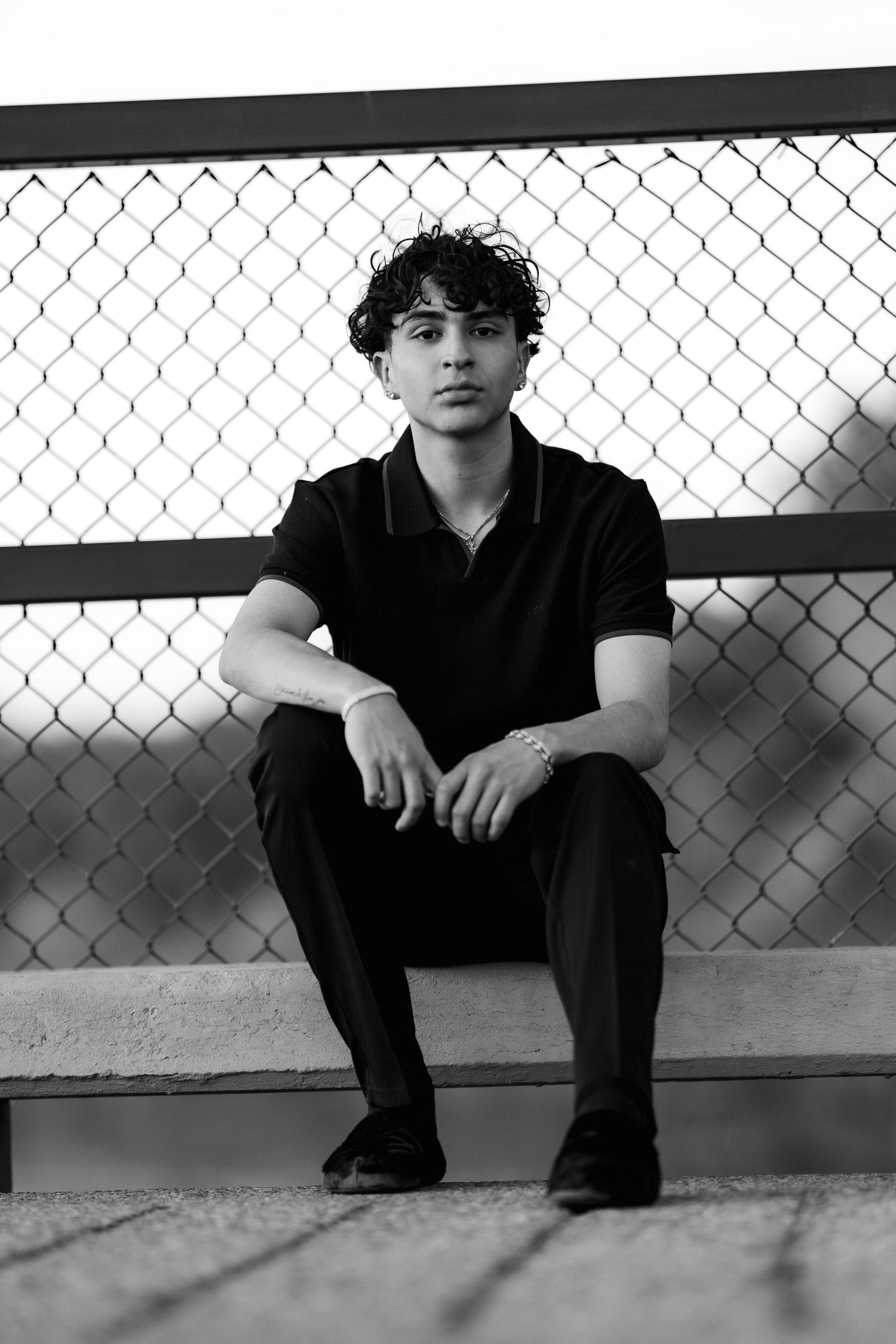 A black and white portrait featuring someone seated on a bench in front of a chain-link fence.