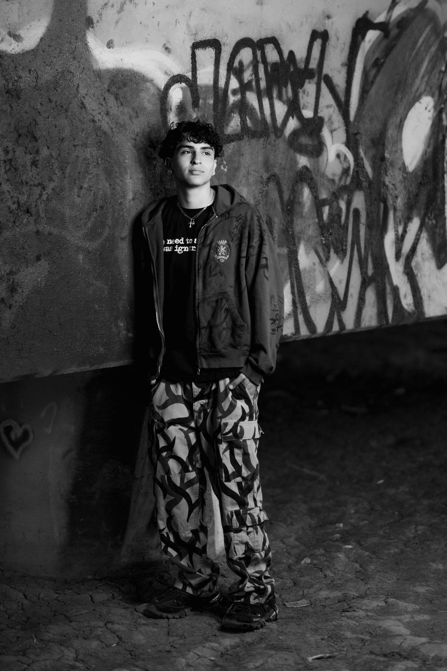 Black and white portrait showing person in camouflage pants and dark jacket standing against graffiti wall.
