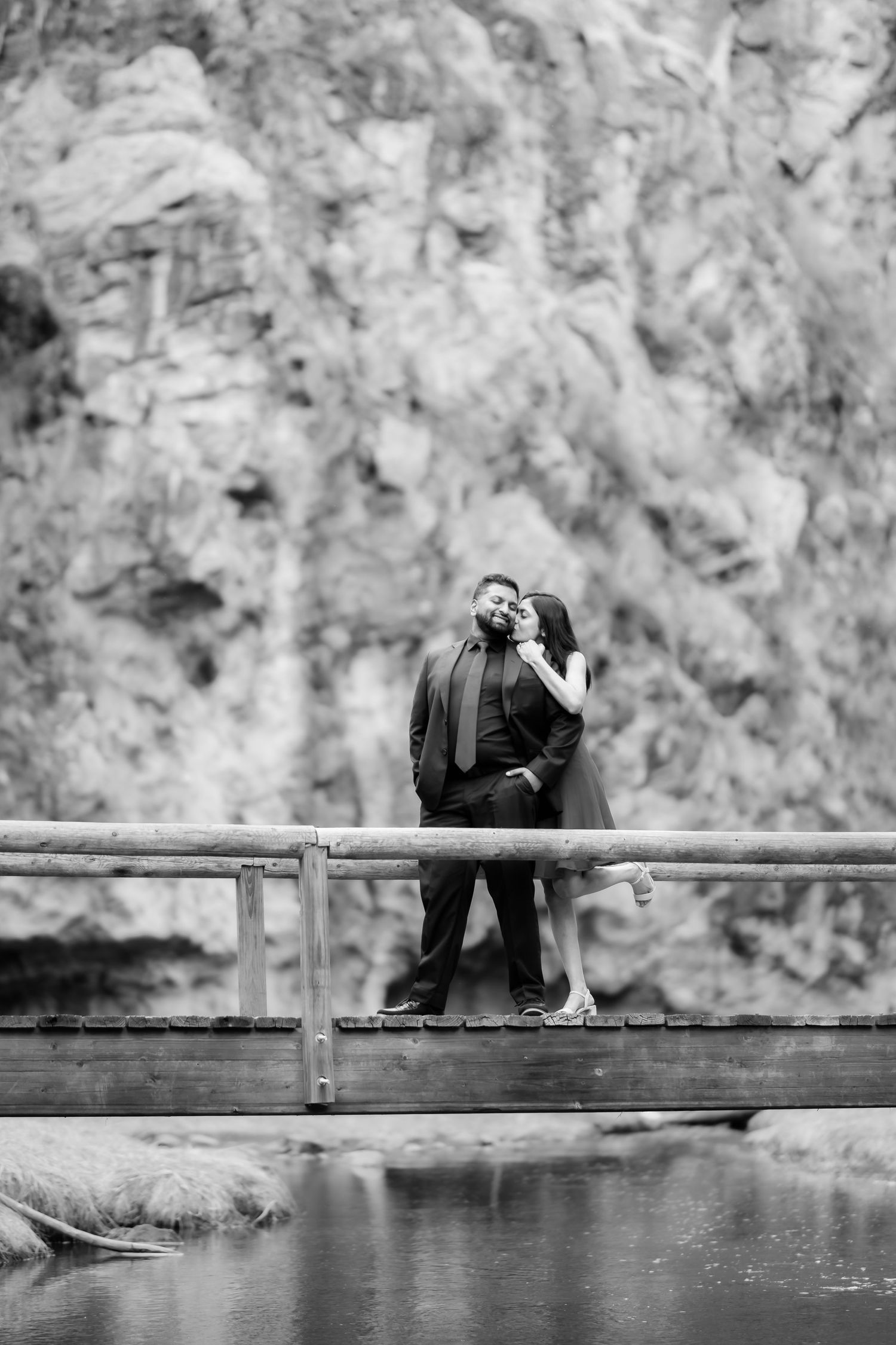 Black and white photo of couple sharing romantic moment on bridge with mountain backdrop.