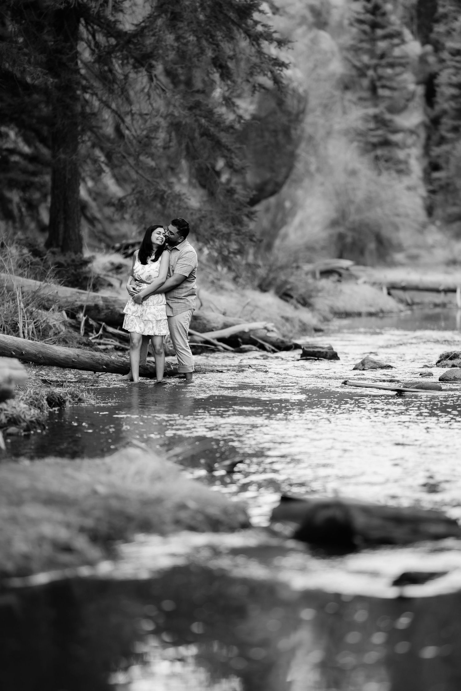 Black and white photo of couple embracing by mountain stream with evergreen forest backdrop.