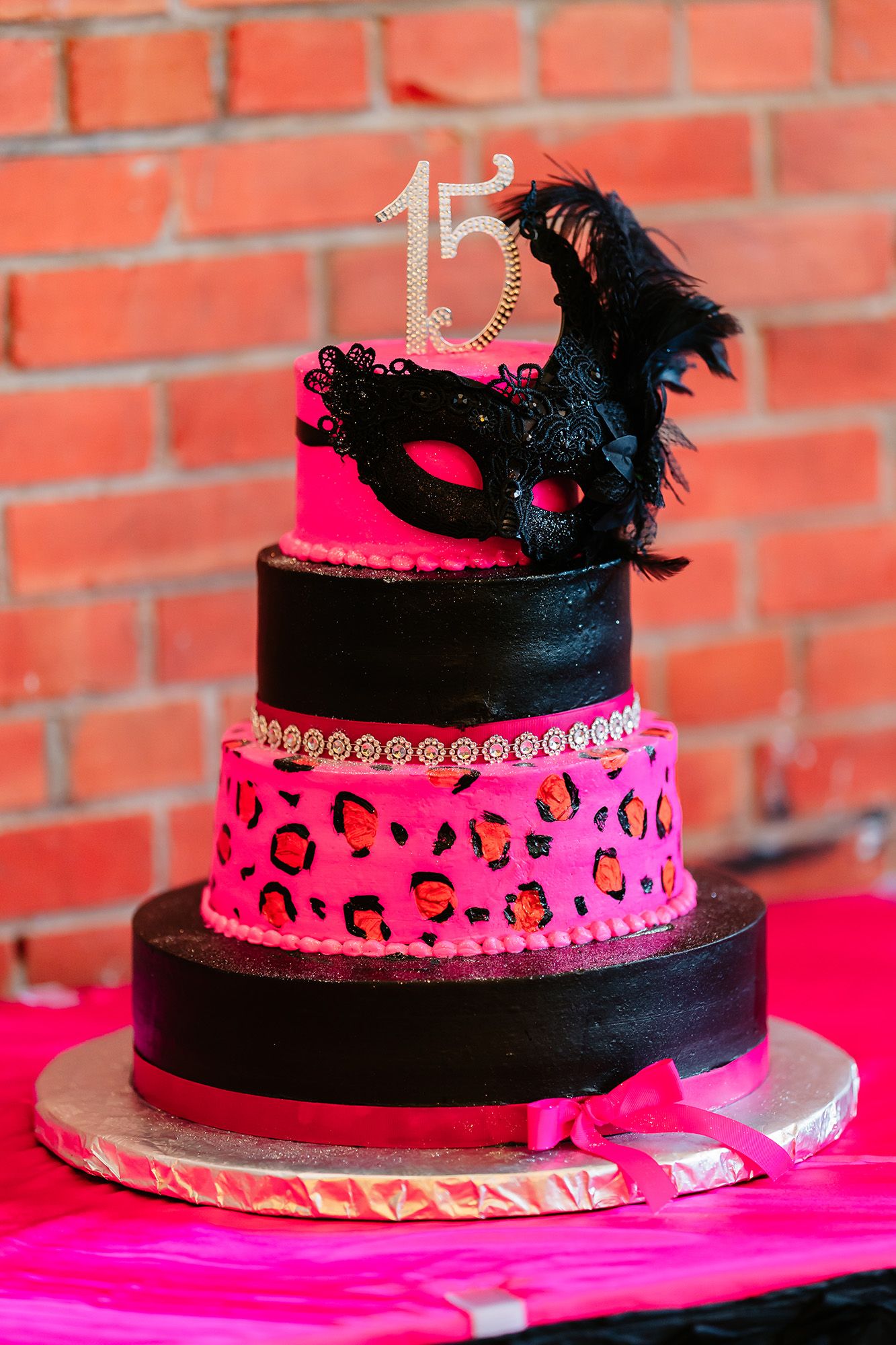 A four-tier masquerade themed birthday cake with hot pink and black layers featuring leopard print and a feathered mask topper.