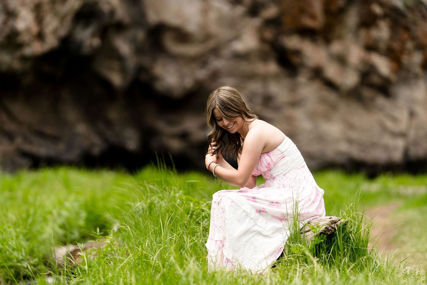 Young woman in pink floral dress kneels in tall green grass near rocky backdrop outdoors.