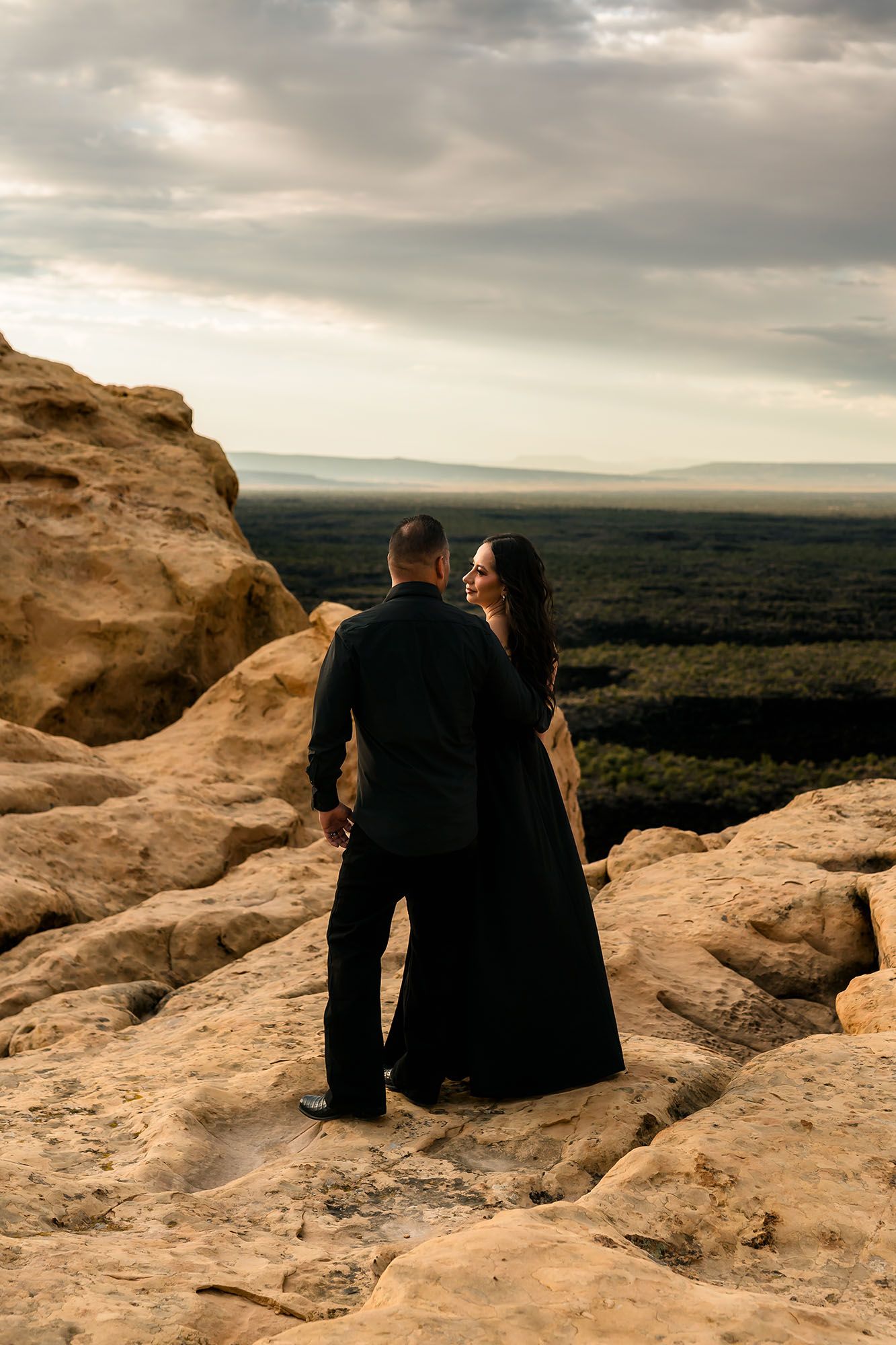 Couple in black outfits standing on rocky cliff overlooking vast forest landscape under cloudy sky.