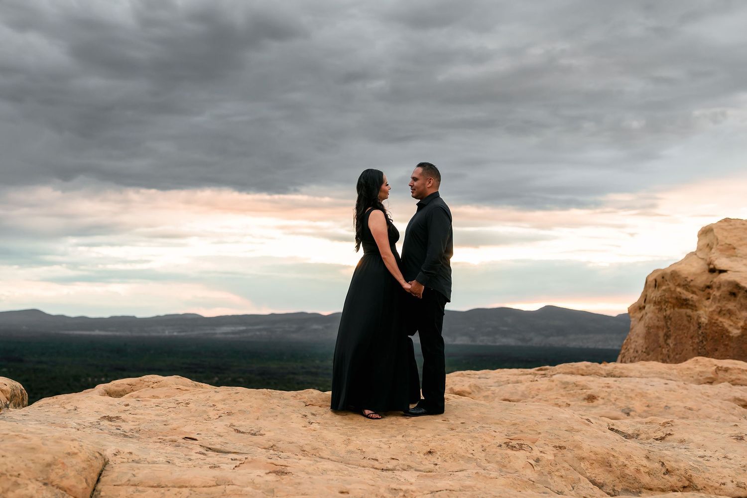 Couple in black attire holding hands on rocky overlook with dramatic cloudy sunset sky and mountain views.