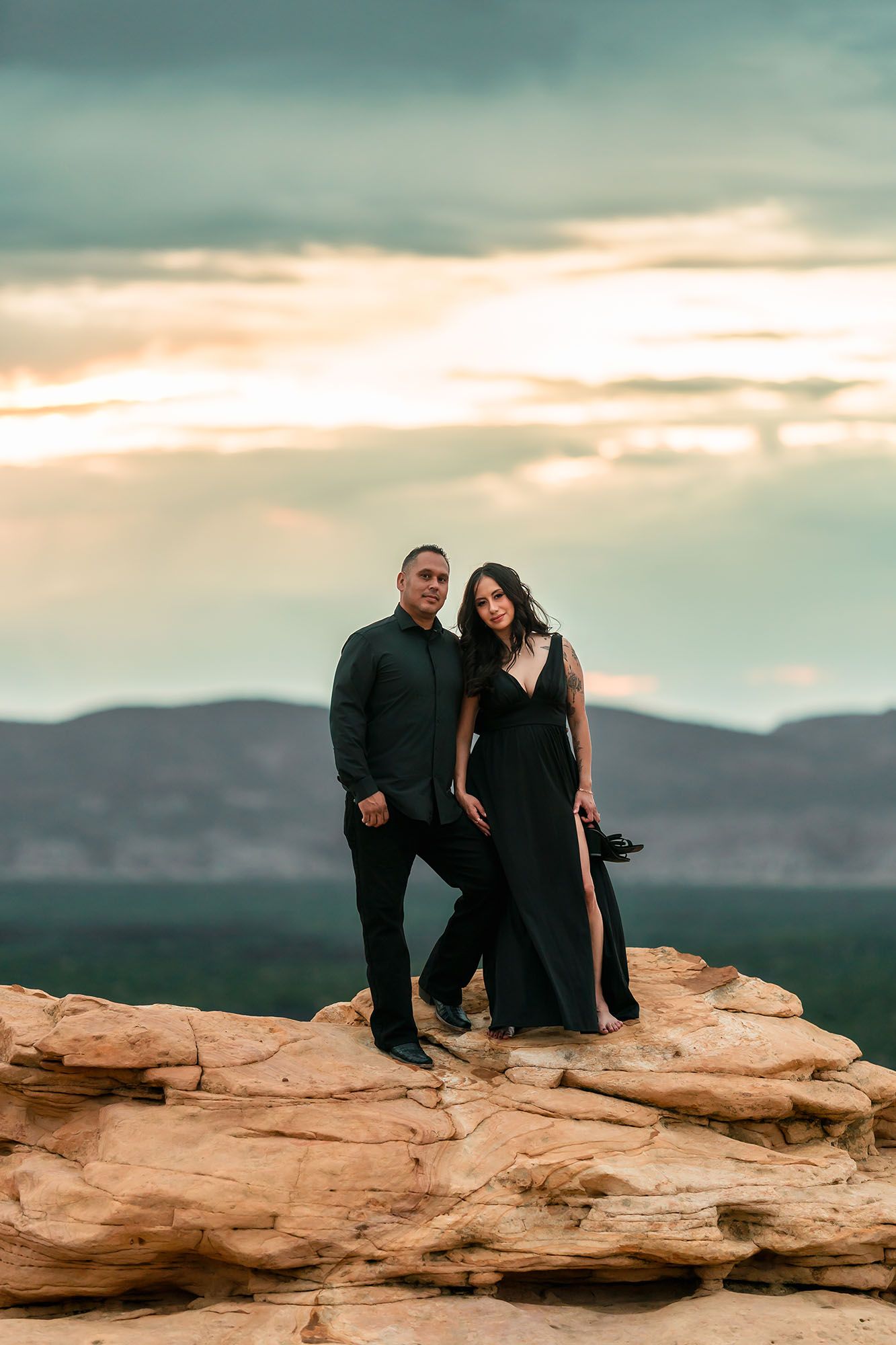 Couple in black outfits posing on red rock formation at sunset with mountain landscape backdrop.