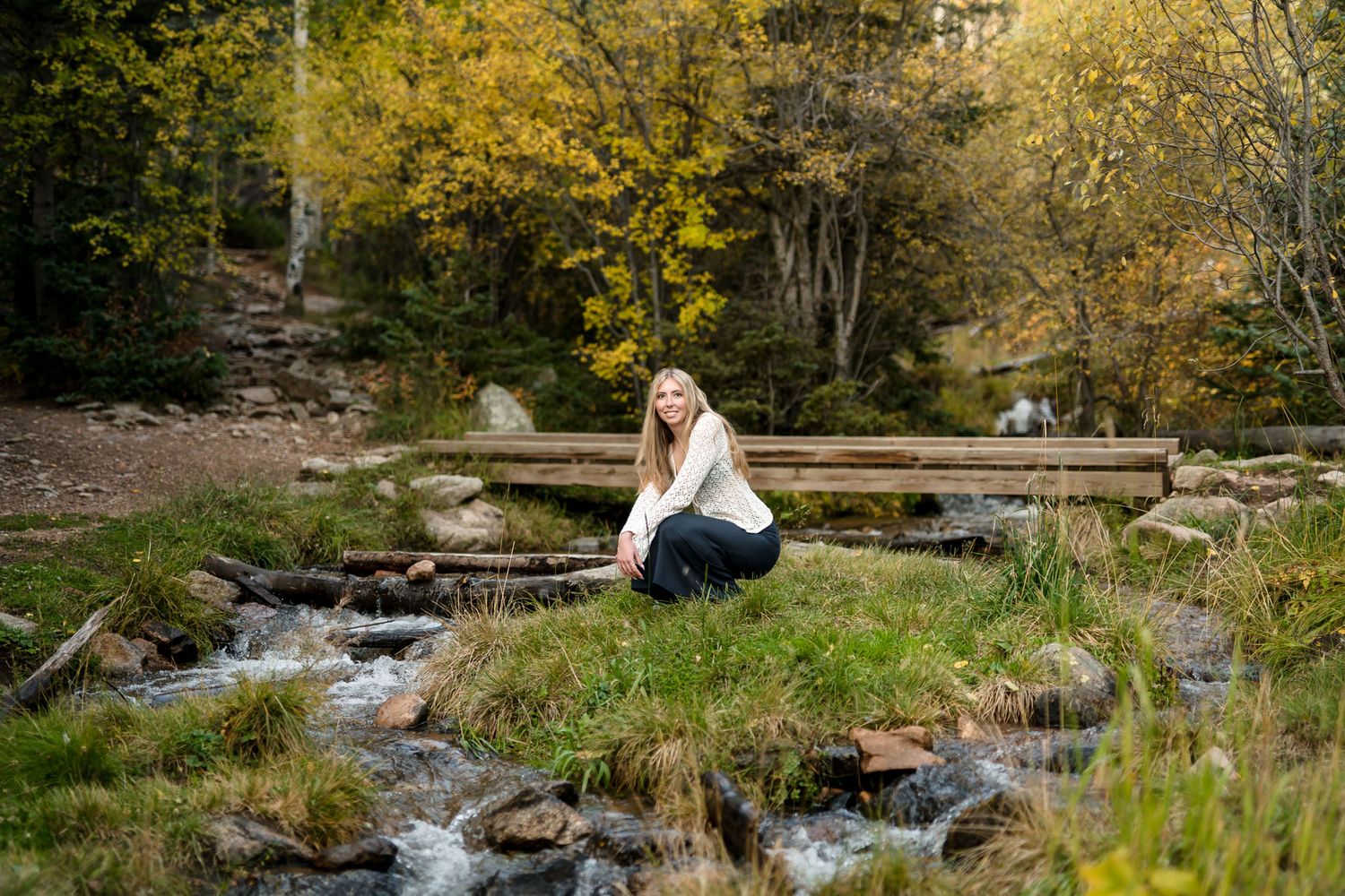 Smiling blonde woman crouches beside a rocky stream with a wooden bridge and fall trees behind her.