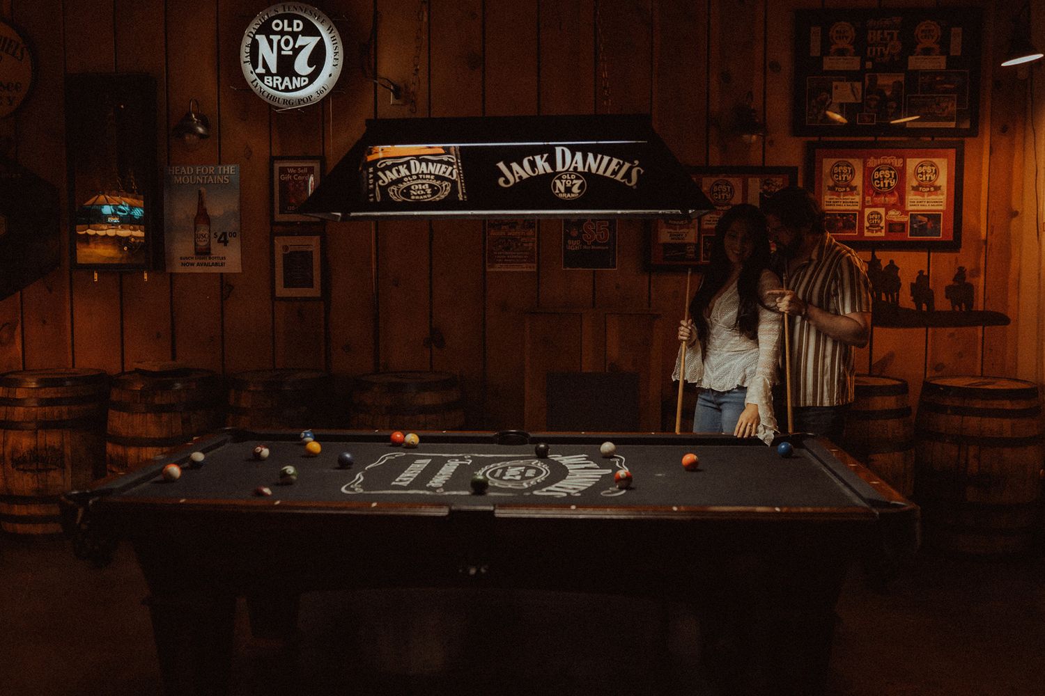 Woman playing pool on a Jack Daniels branded billiard table in a rustic bar with whiskey barrel decor.