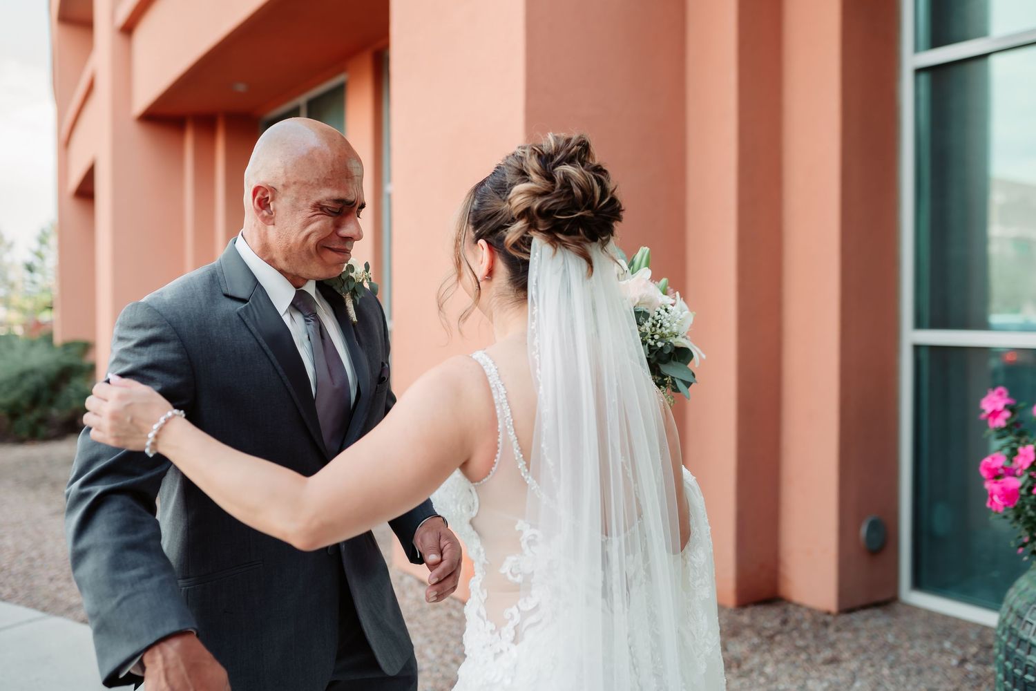 A bride in a white wedding gown with veil shares a tender dance moment outside a coral-colored building.